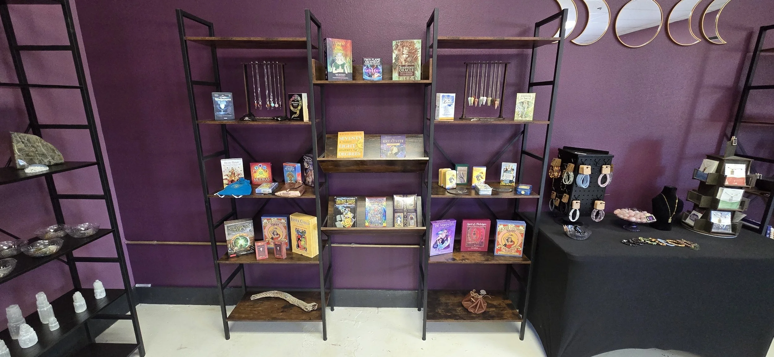 A display of various witchcraft and spiritual items, including jewelry, tarot cards, crystals, and books, against a purple wall.