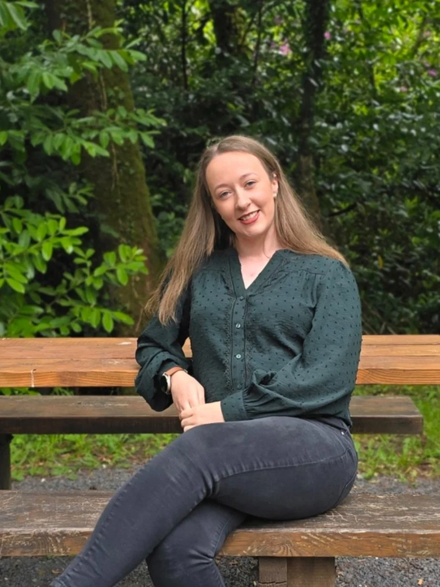 A young woman with long hair sitting on a wooden bench outdoors in a green, wooded area, smiling at the camera.