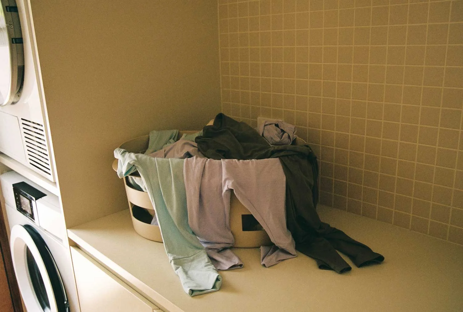 A laundry room counter with a basket of clothes and laundry spilling over, laundry machines on the side, and a tiled wall in the background.