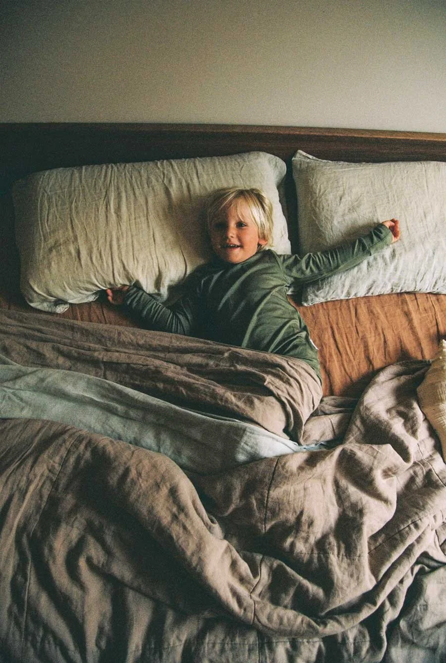 A young child lying on a bed with large pillows, smiling and wearing a green long-sleeve outfit, surrounded by rumpled bedding.