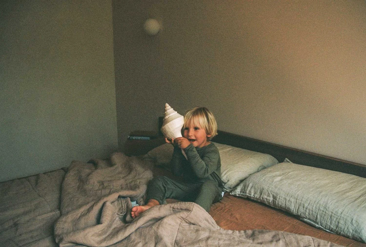 A young boy with blonde hair sitting on a bed, holding a seashell shaped object near his face, with pillows and blankets around him in a dimly lit room.