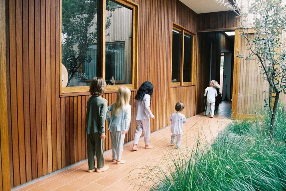 Five children standing on a wooden porch outside a house with wooden siding, greenery in the foreground, and an open door leading back inside.