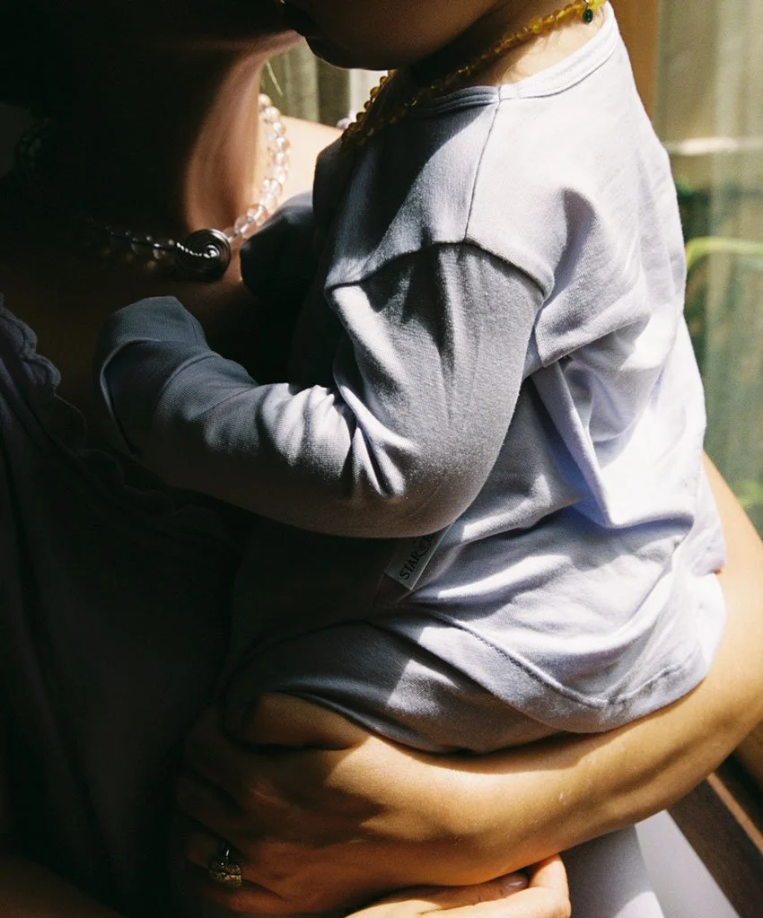 A woman holding a small child, with both their faces partially visible. The woman is wearing a pearl necklace and a black shirt, while the child is dressed in a gray long-sleeve shirt and shorts, sitting on the woman's lap.