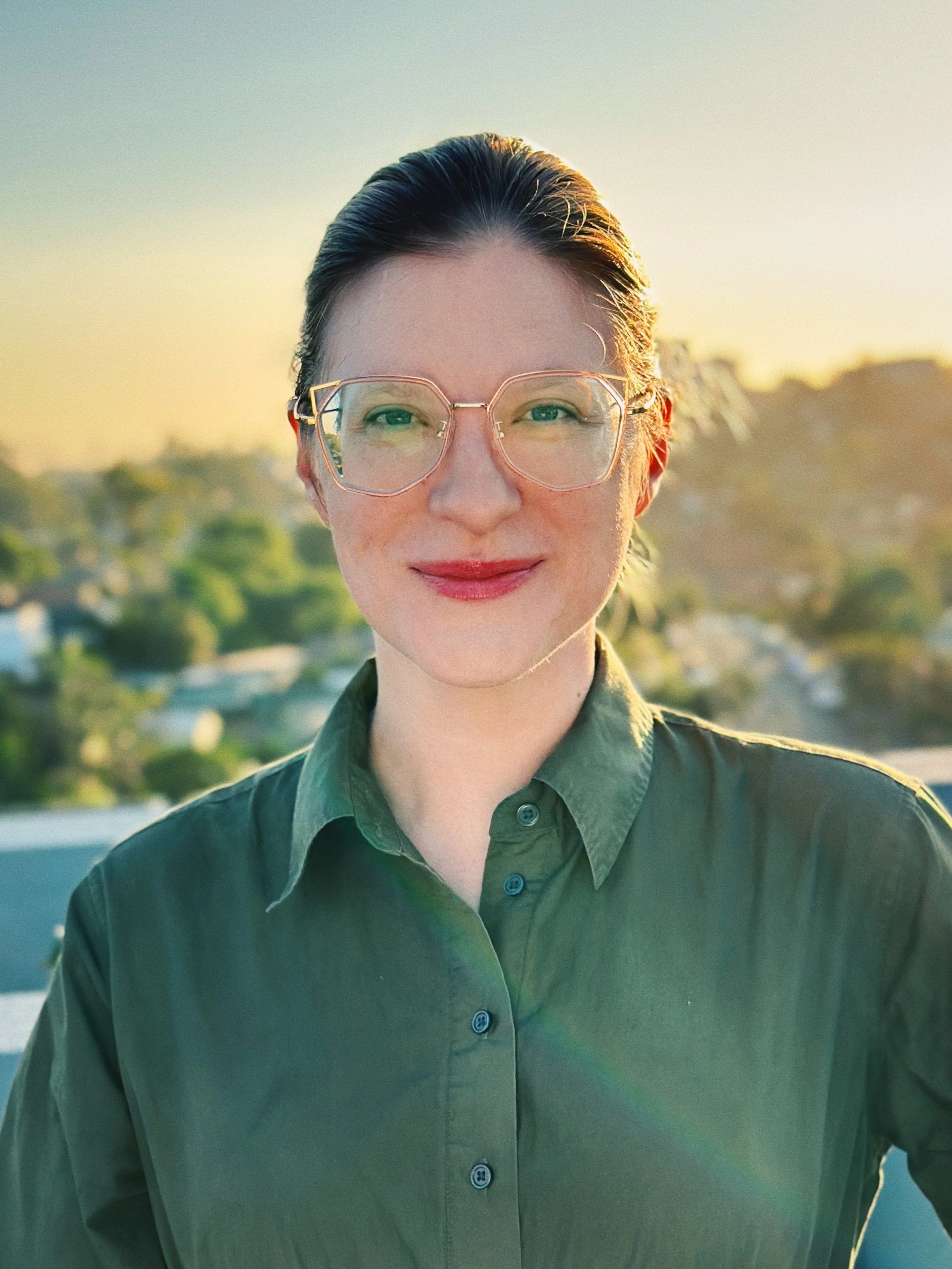 A woman with tied back hair and glasses smiling outdoors during sunrise, wearing a green button-up shirt.