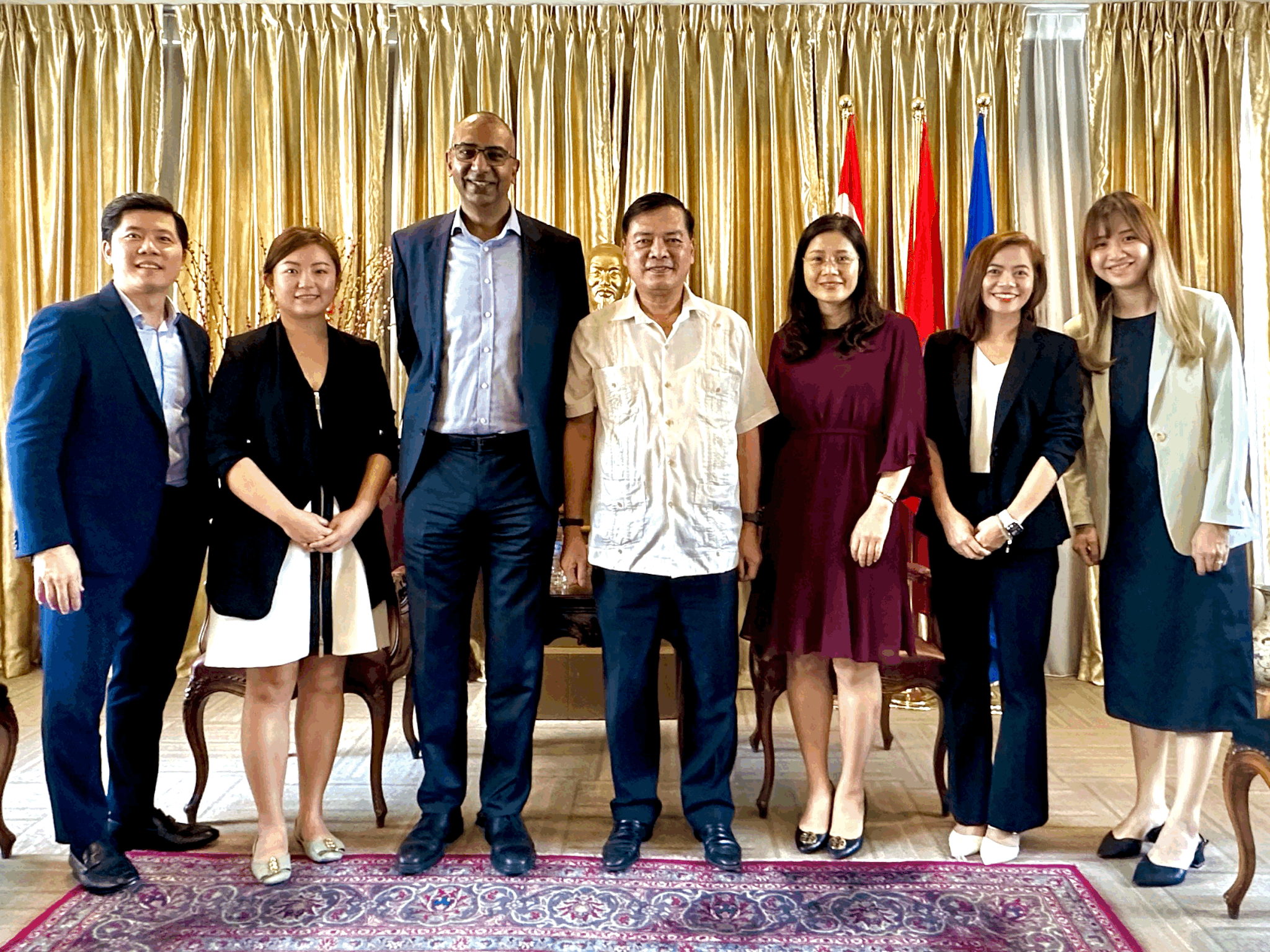Group of eight diverse people standing together in formal attire in a room with gold curtains and flags in the background.