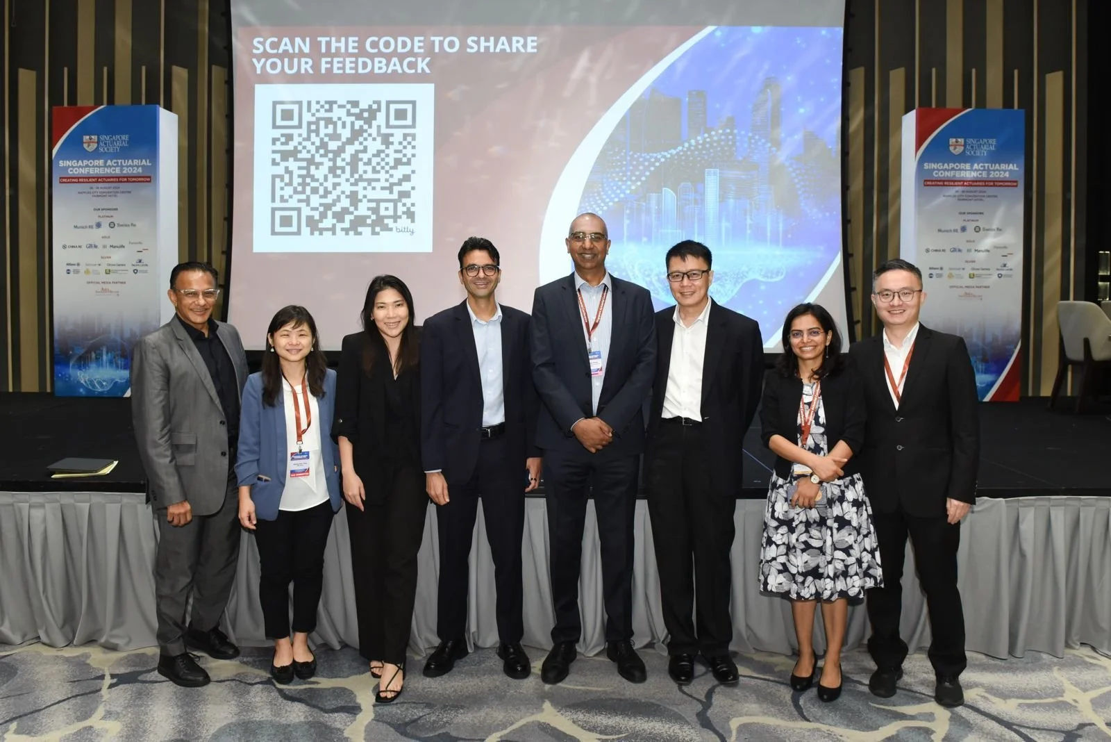 Group of nine diverse professionals at the Singapore Actuarial Conference 2024, standing on stage in front of a large screen with a QR code and conference info.