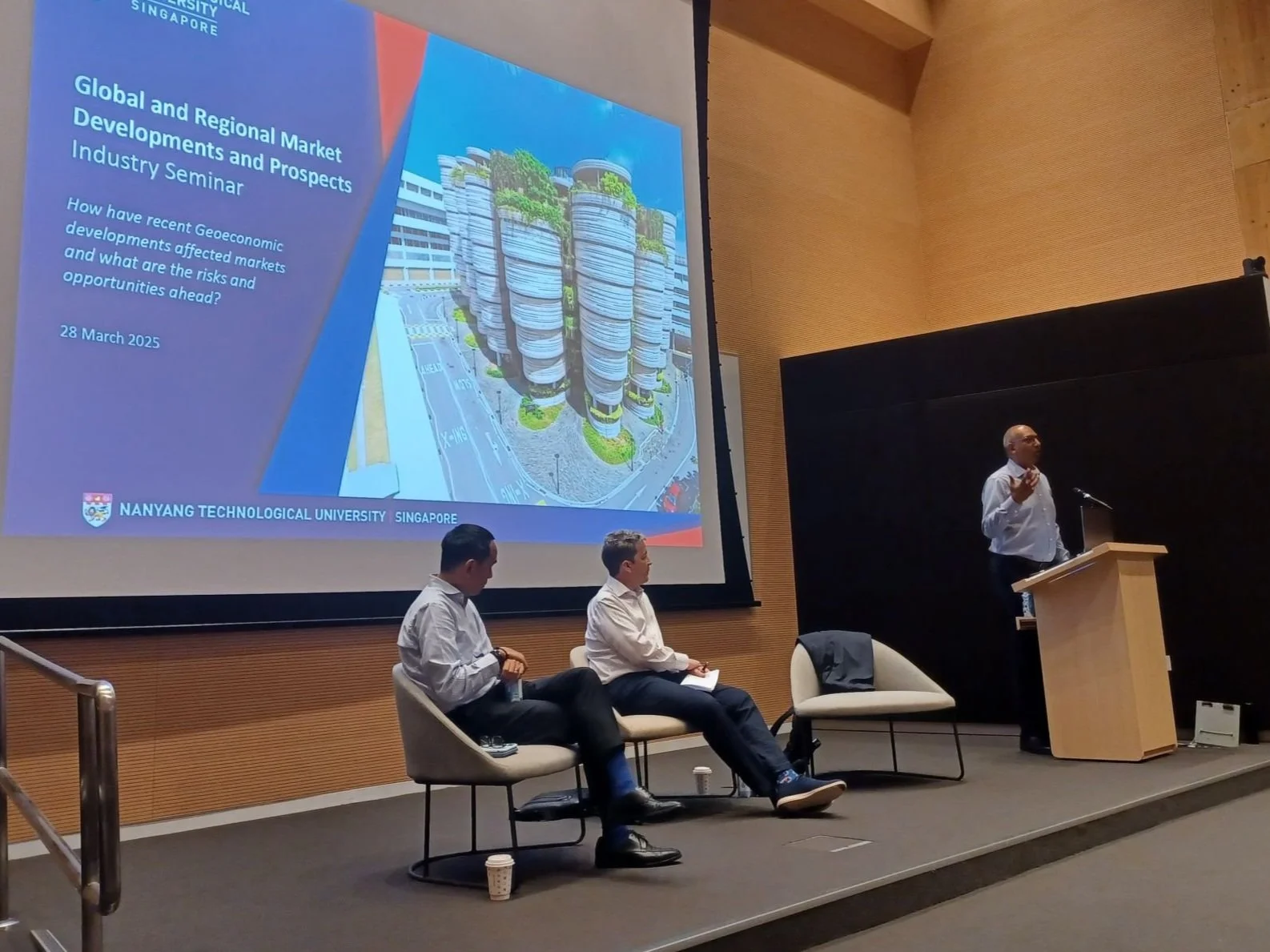 A person giving a presentation at a seminar about global and regional market developments at Nanyang Technological University Singapore. There are two individuals seated on stage and a slide projected on a large screen showing the seminar title, date, and an architectural rendering of a building complex.
