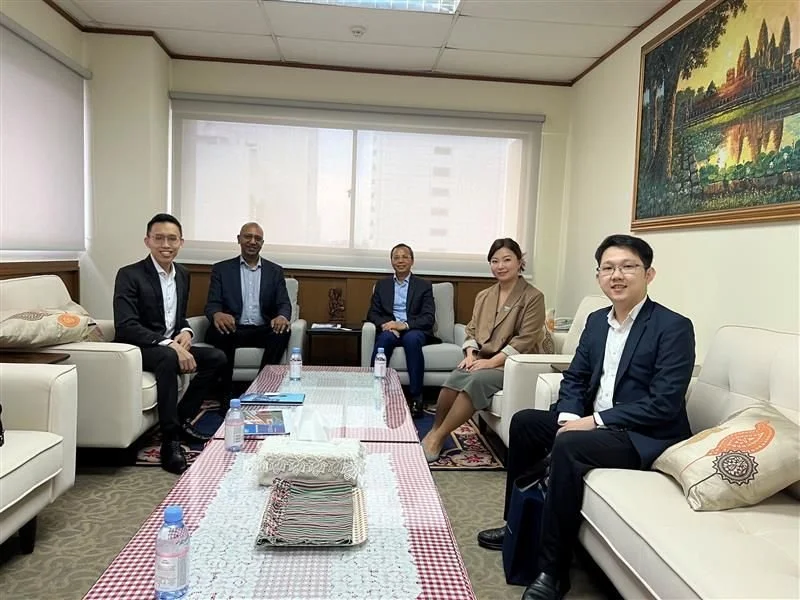 Five people sitting in a meeting room with sofas, a table with bottled water and documents, a large window, and a painting on the wall.