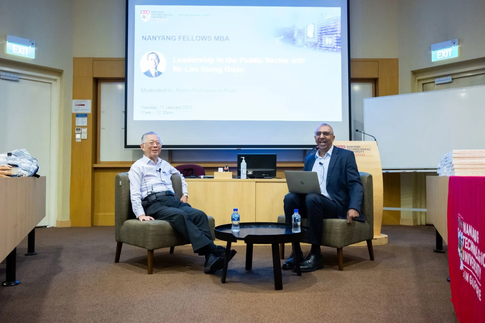 Two men seated on armchairs in a conference room, smiling. A large screen behind them displays a presentation titled 'Leadership in the Public Sector with Mr. Lim Siong Guan,' moderated by Professor Youngesh Khattri, at Nanyang Technological University. There are water bottles on a small round table between them, and a laptop on one man's lap.
