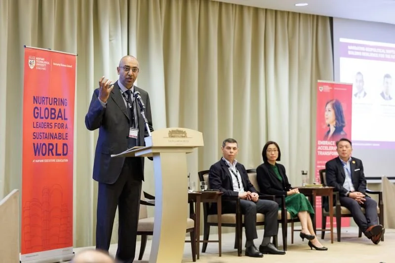 A man in a suit speaking at a podium during a conference, with three seated panelists and two banners in the background promoting global leadership and sustainability.