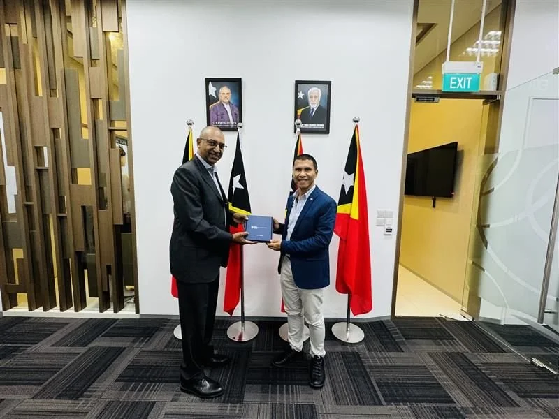 Two men in suits exchanging a document in an office with two flags, two portraits on the wall, and an exit sign.