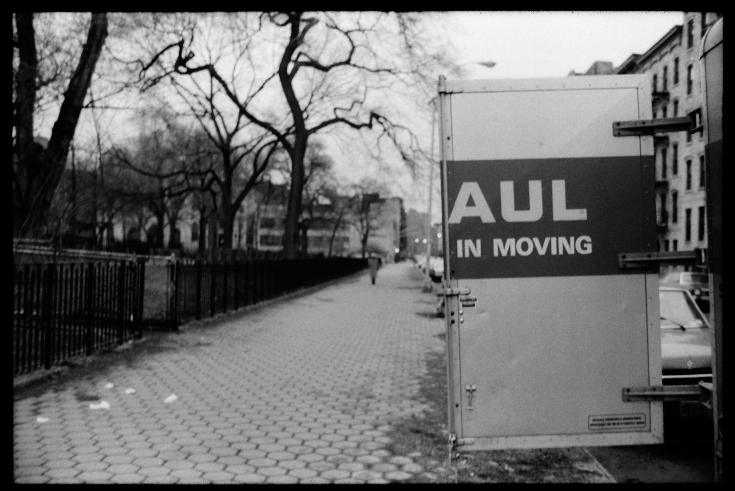 Black and white photo of a sidewalk with leafless trees, a fence on the left, and buildings in the background. A sign on a U-Haul box trailer on the right reads 'AUL IN MOVING'.