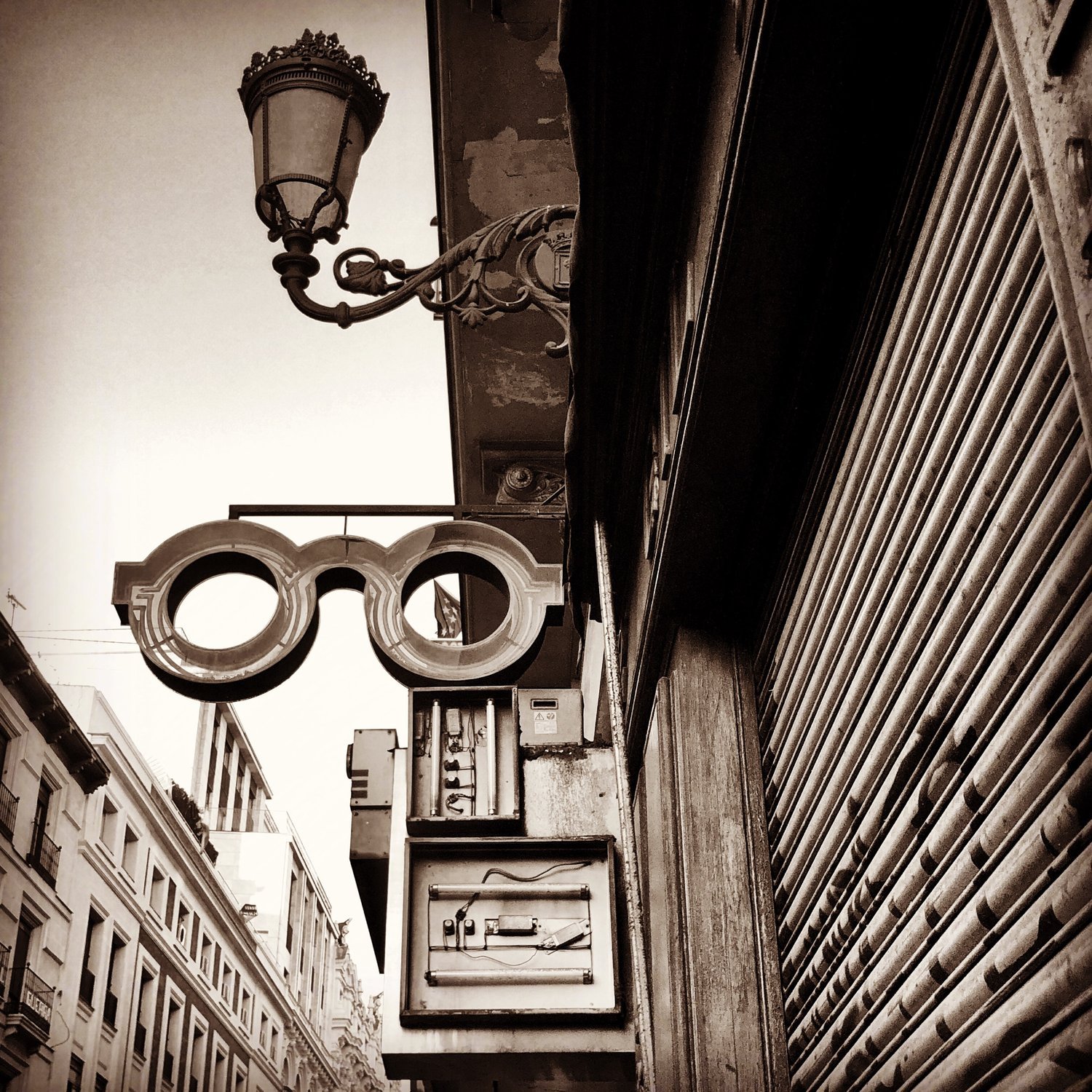 Street view with vintage-style street lamp and a neon sign for glasses or eyewear store, featuring electrical equipment and a roll-up metal shutter.