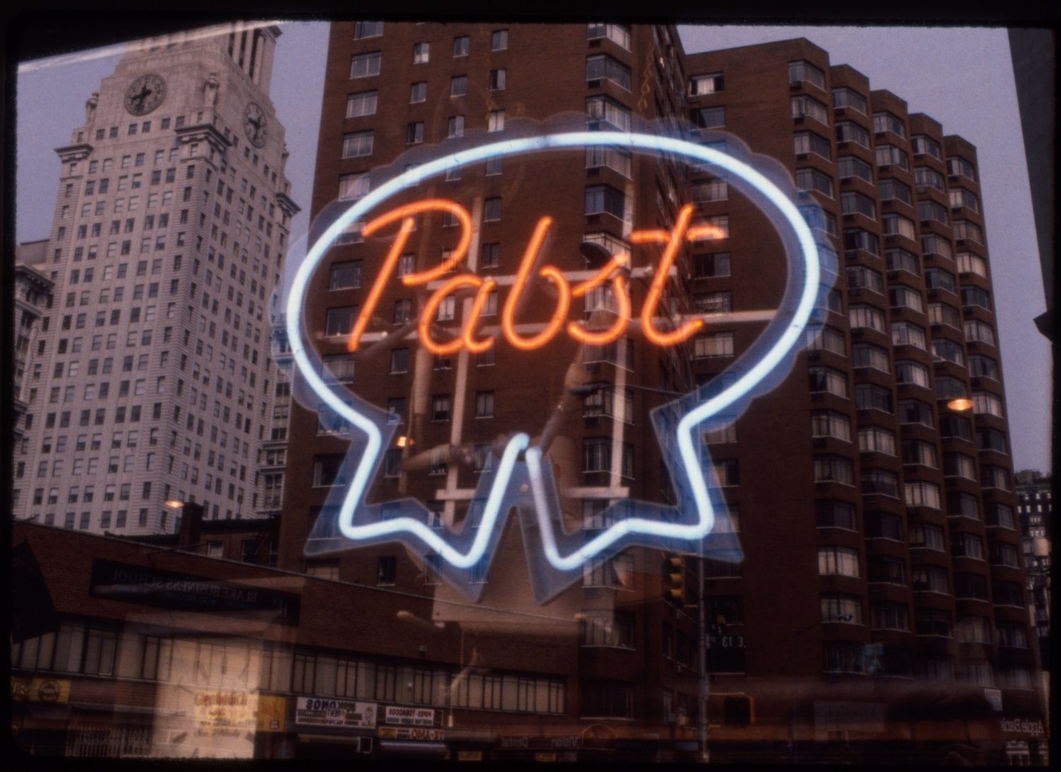 Neon sign in the shape of a speech bubble with the word 'Pabst' written inside, reflecting city buildings on a window.