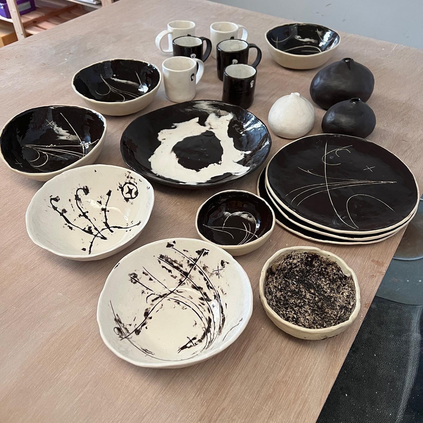 Black and white ceramic bowls, plates, and mugs with abstract designs arranged on a wooden table.