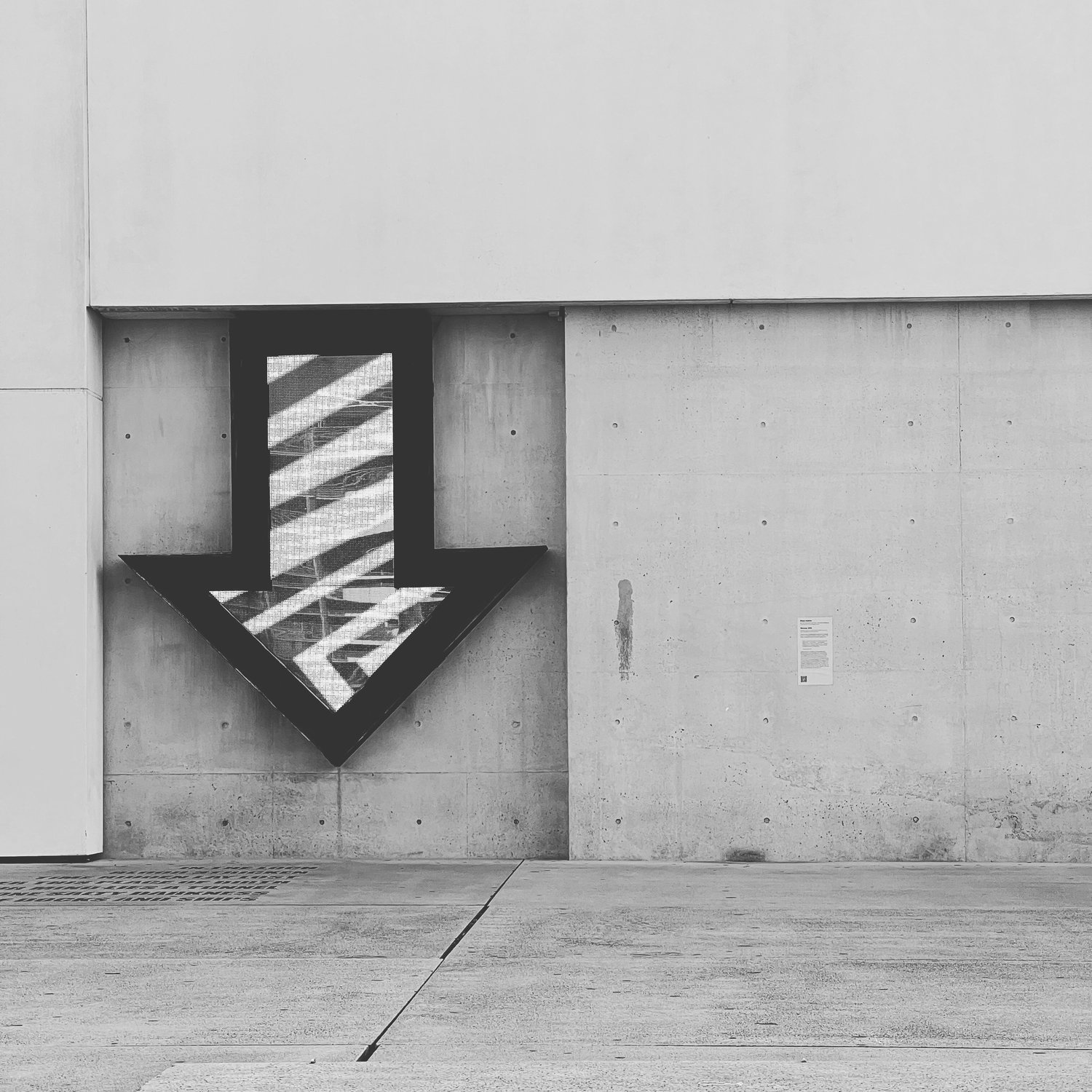 Black and white photo of an arrow sign pointing downward placed on a concrete wall.