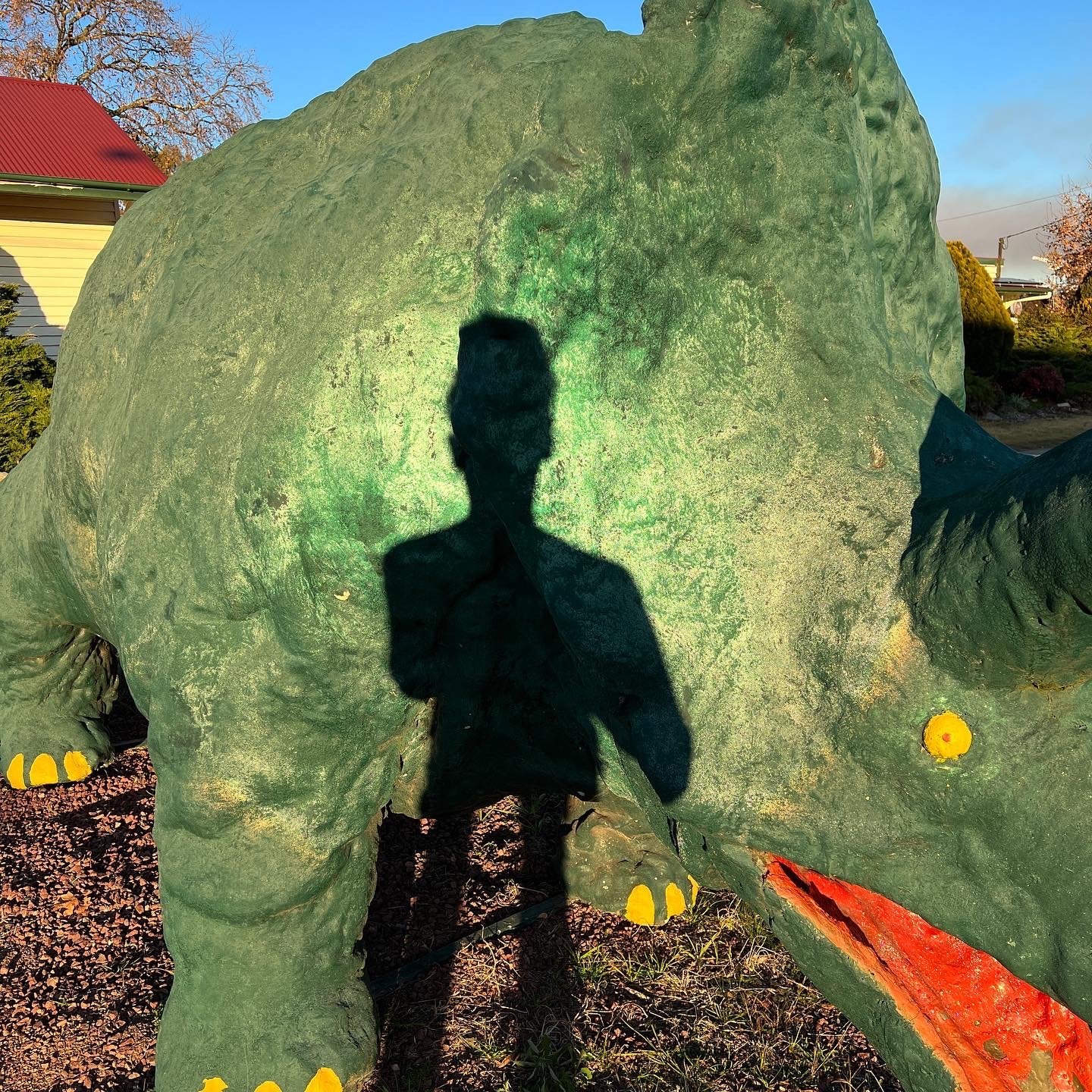 Shadow of a person taking a photo, cast onto a large green sculpture of a dinosaur, outdoors with a house and trees in the background.