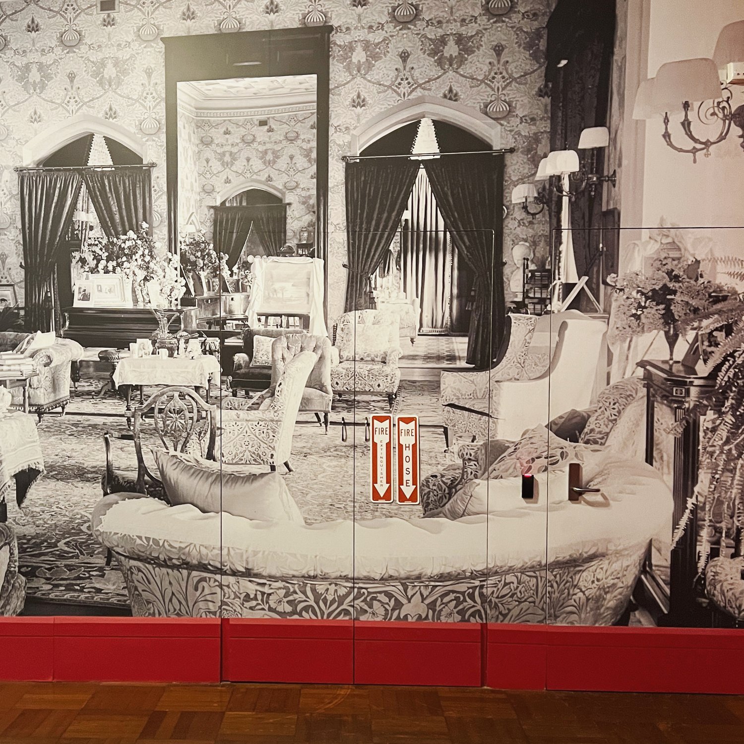 A black-and-white photograph of an elegantly furnished vintage living room with ornate wallpaper, several upholstered chairs and sofas, a fireplace, and a large mirror. There are floral arrangements, lamps, and a fire hazard sign in the center.