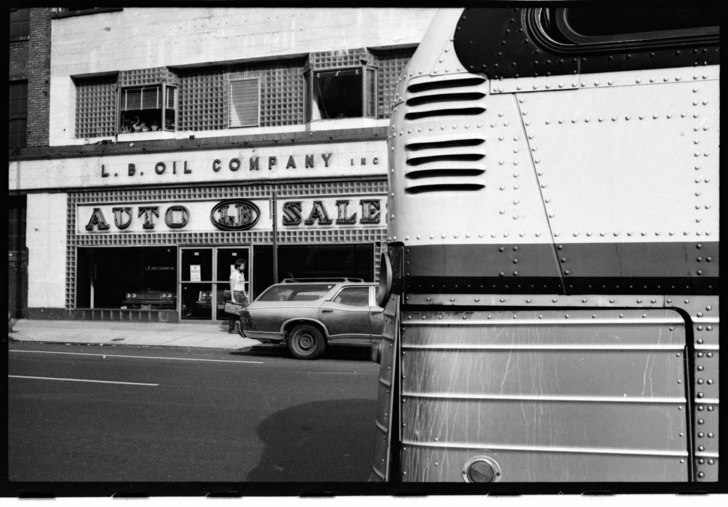 A black and white photo of an urban street scene featuring a storefront sign that reads 'L.B. Oil Company Inc.' and 'Auto Sale.' There is a parked car in front of the store and a woman walking on the sidewalk. A large vehicle or trailer is partially