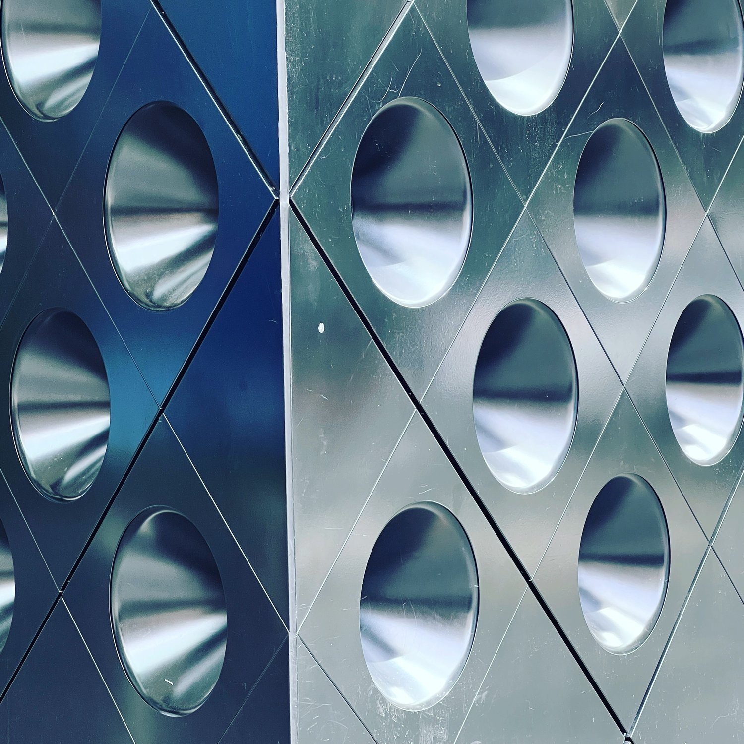 Close-up of a metallic structure with multiple large circular holes, reflecting light and surrounding environment.