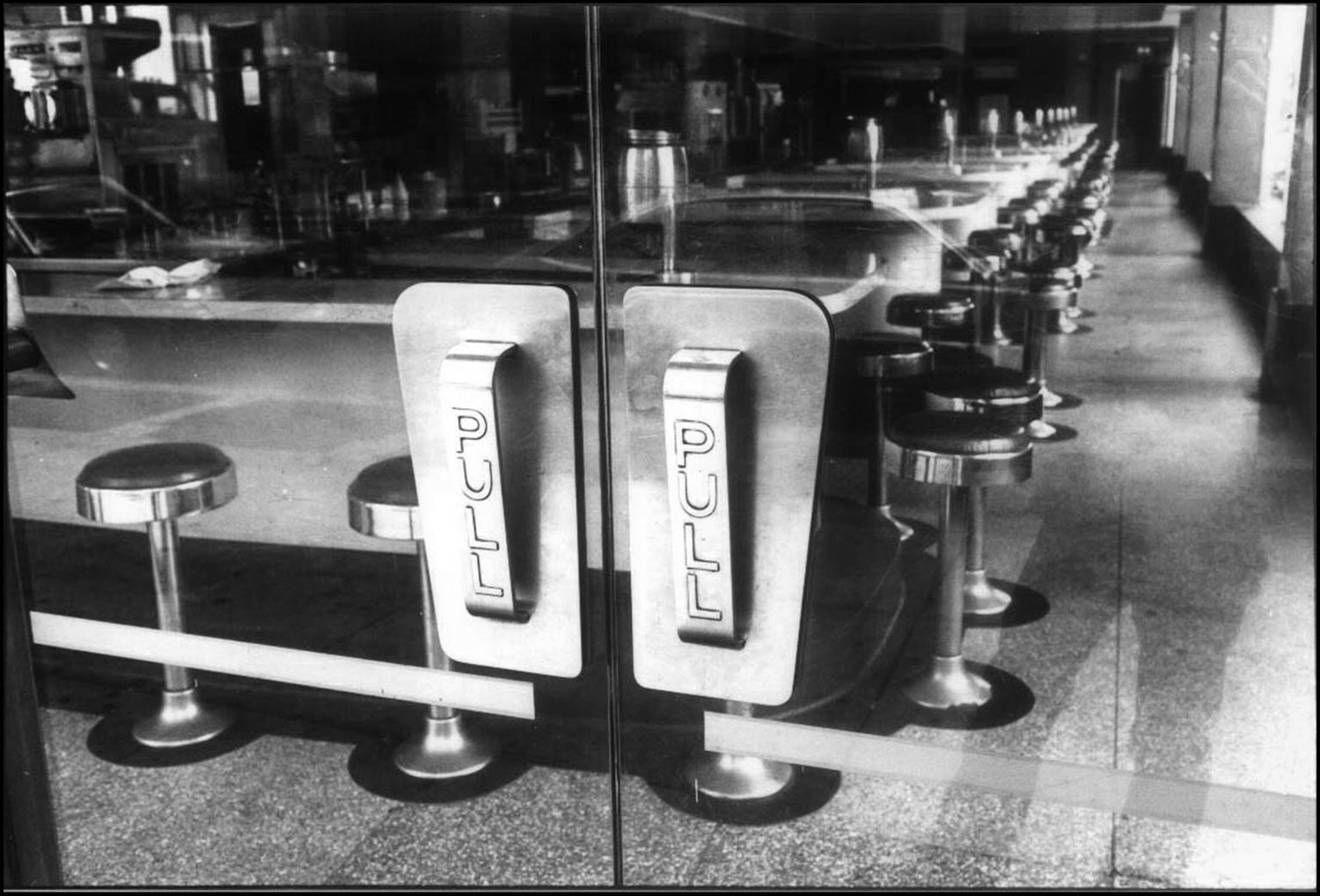 Black and white photo of a diner with a row of stools along a counter, viewed through glass doors with 'PUSH' handles.