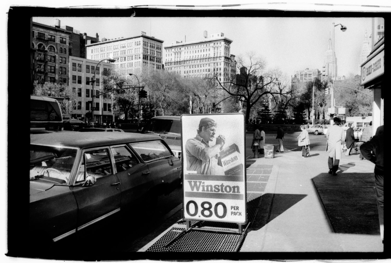 Black and white photo of a city sidewalk with pedestrians, parked cars, and a billboard advertising Winston cigarettes for 0.80 per pack, with a background of tall buildings, trees, and a street.