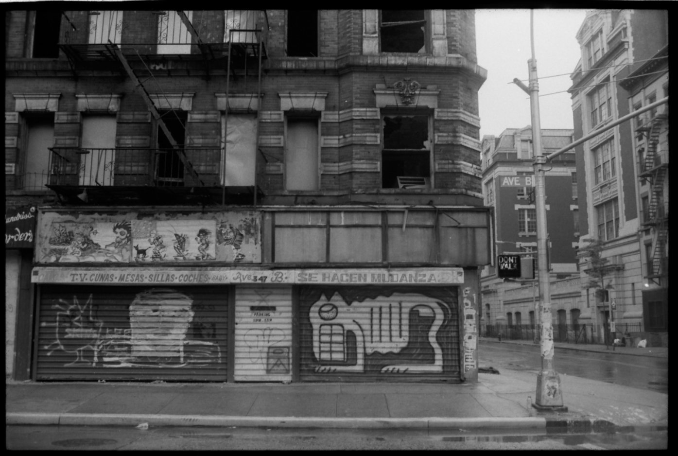 Black and white photo of an abandoned building on a city street corner with graffiti on the shutter and a sign that says 'T.V. CUNAS MESAS SILLAS COCHES' and a graffiti drawing of a kitten and a hand making a peace sign on the shutter. A street sign