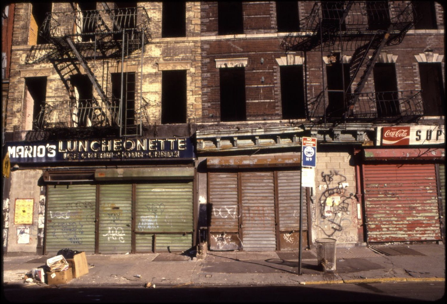 A brown brick building with boarded-up windows and graffiti on the wall. The storefront has a sign that reads 'Mario's Luncheonette' offering ice cream, soda, and candy. The storefront shutters are closed and graffiti covers parts of the building and
