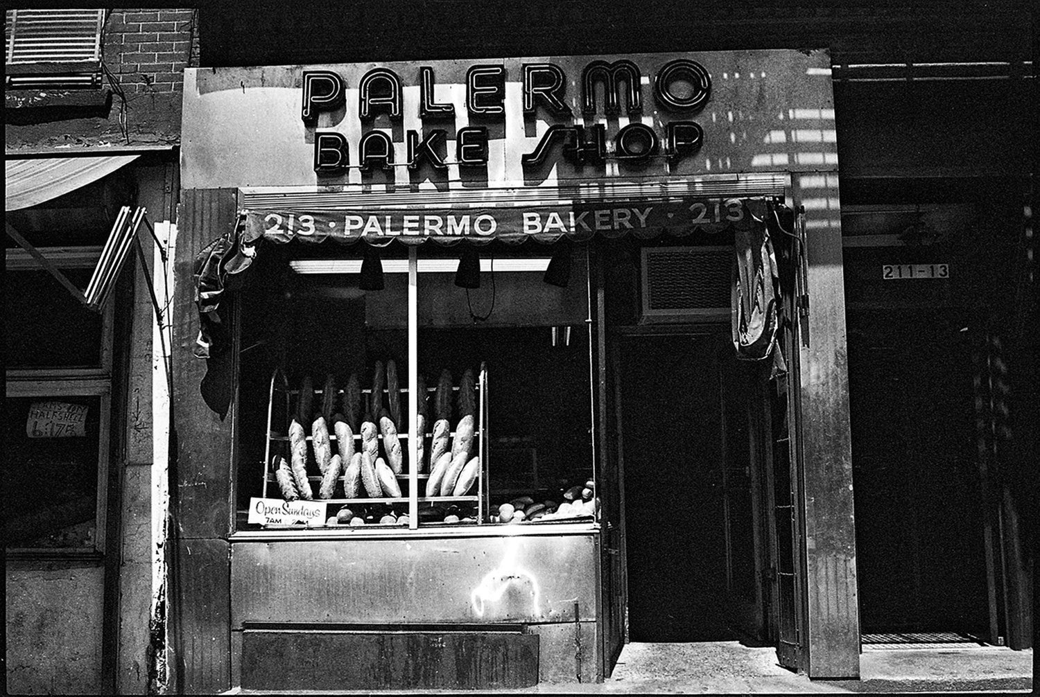 Black and white photo of a bakery storefront with a neon sign reading 'Palermo Bakery' and a display window with bread and hot dogs.