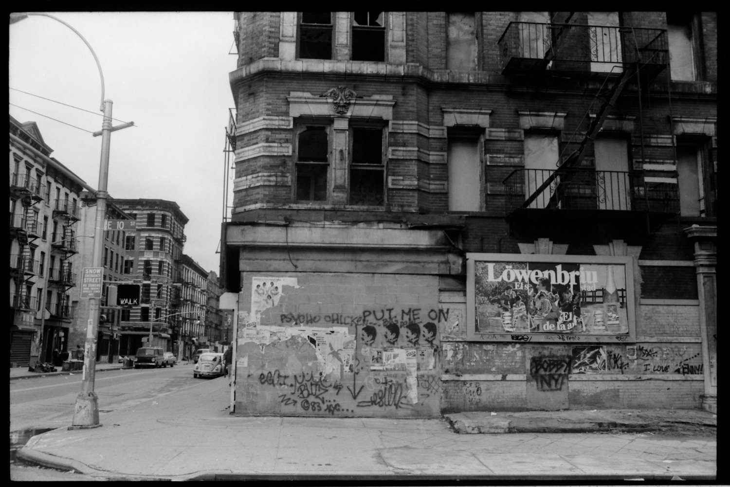 Black and white street scene showing an old brick building with graffiti and posters on the bottom wall, a street with parked cars, and a sign for Snow Street and E 10th Street.