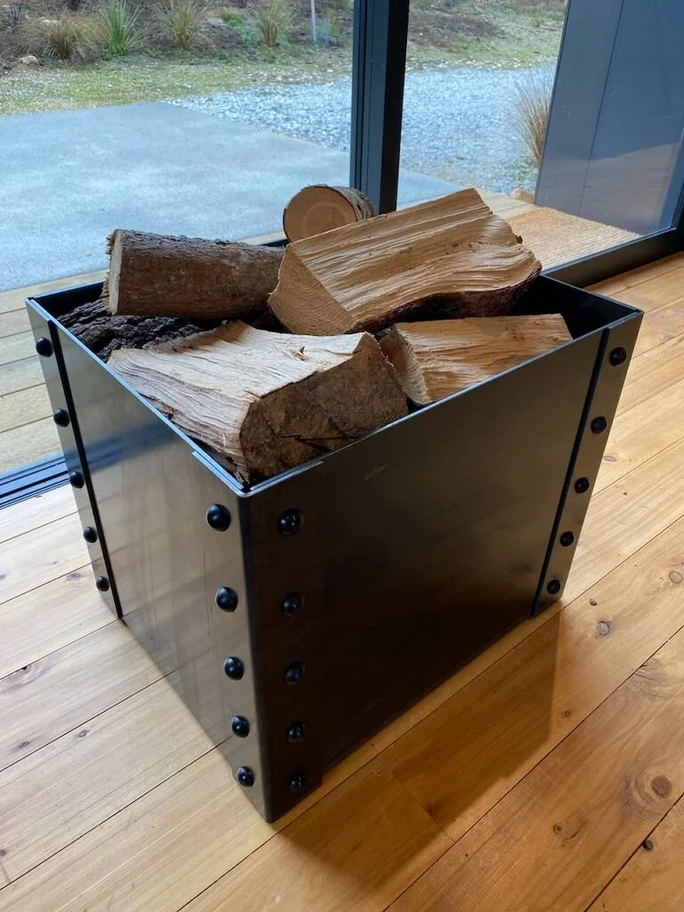 A black metal firewood holder filled with chopped logs, placed on a wooden floor near a glass wall.