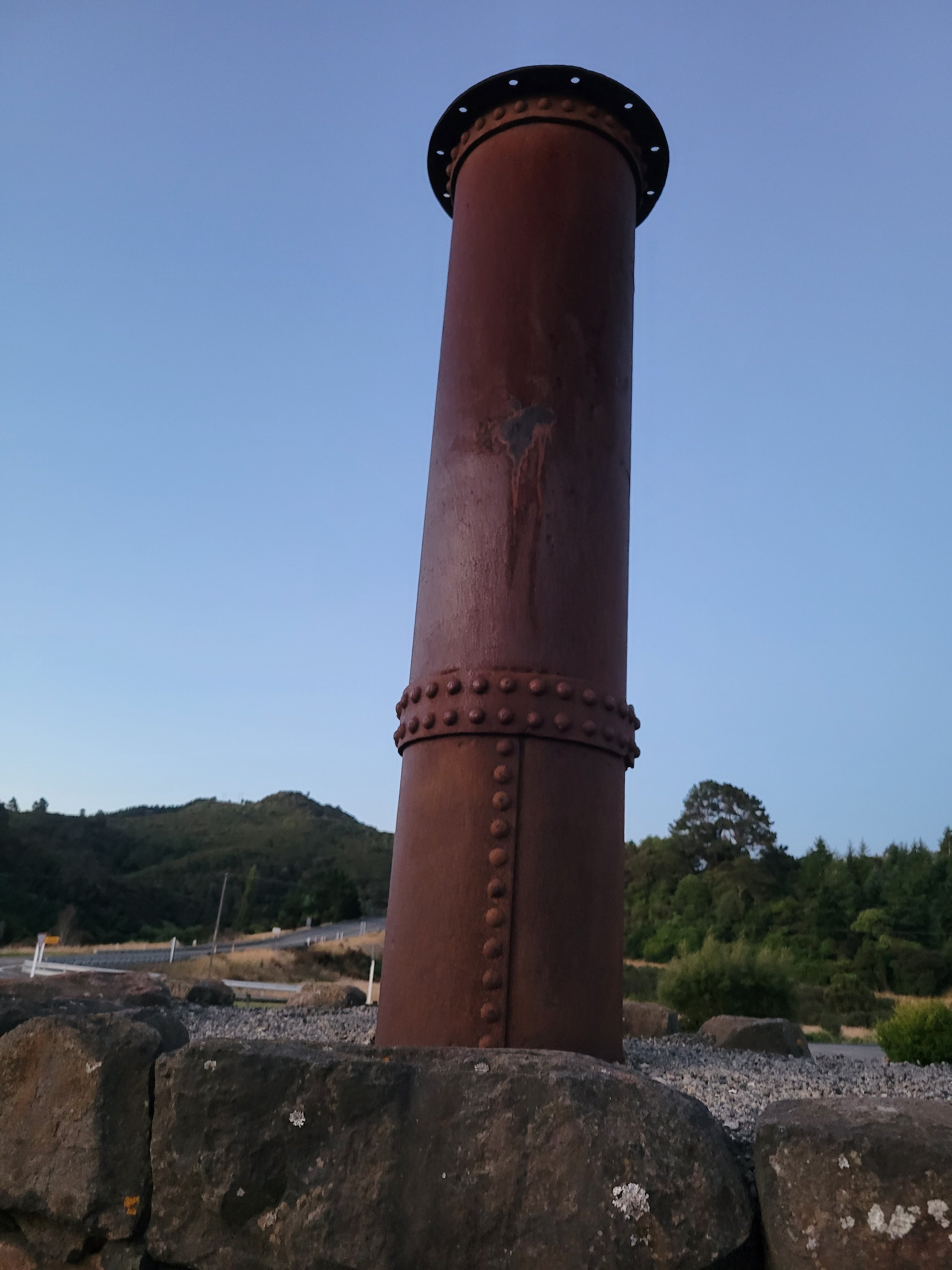 A tall, rusty metal pipe structure standing on a stone base with a rural landscape and hills in the background during dusk.