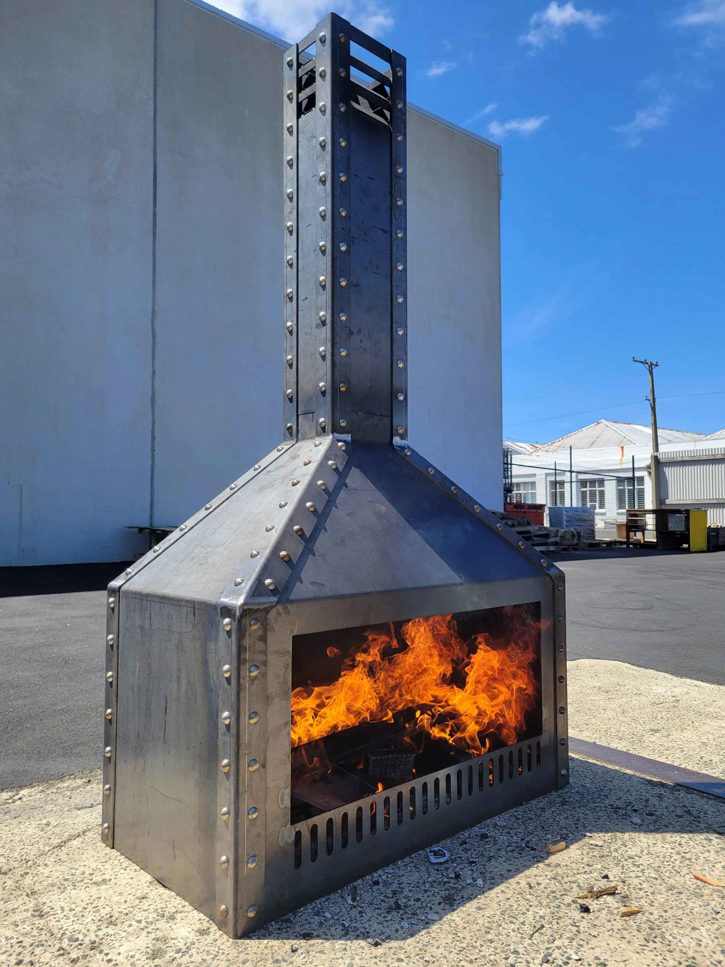 A metal outdoor fireplace with a fire burning inside, positioned on a concrete surface against a light-colored wall with a blue sky in the background.