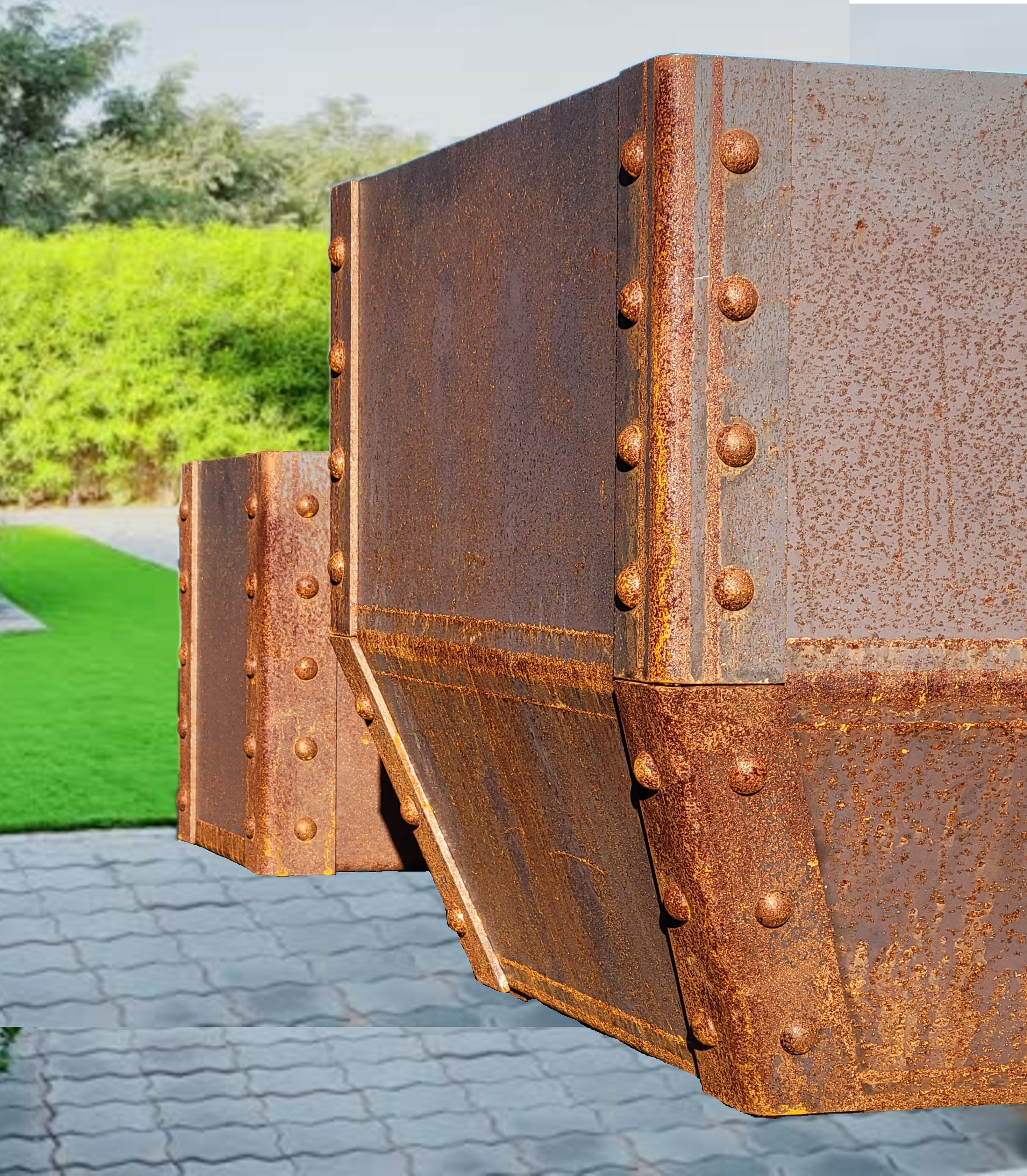 Close-up of rusted metal structure with bolts, outdoors on pavement with grass and trees in background.