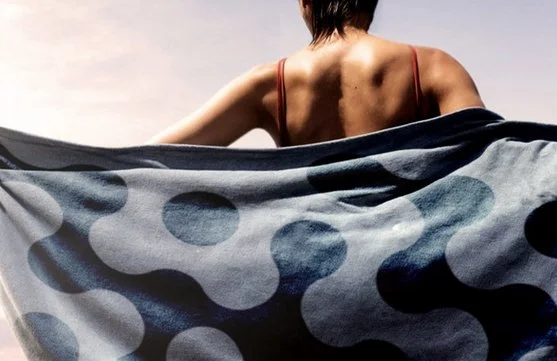 Back of a person with short dark hair wearing a red spaghetti strap top, holding a large polka-dotted fabric in front of them.