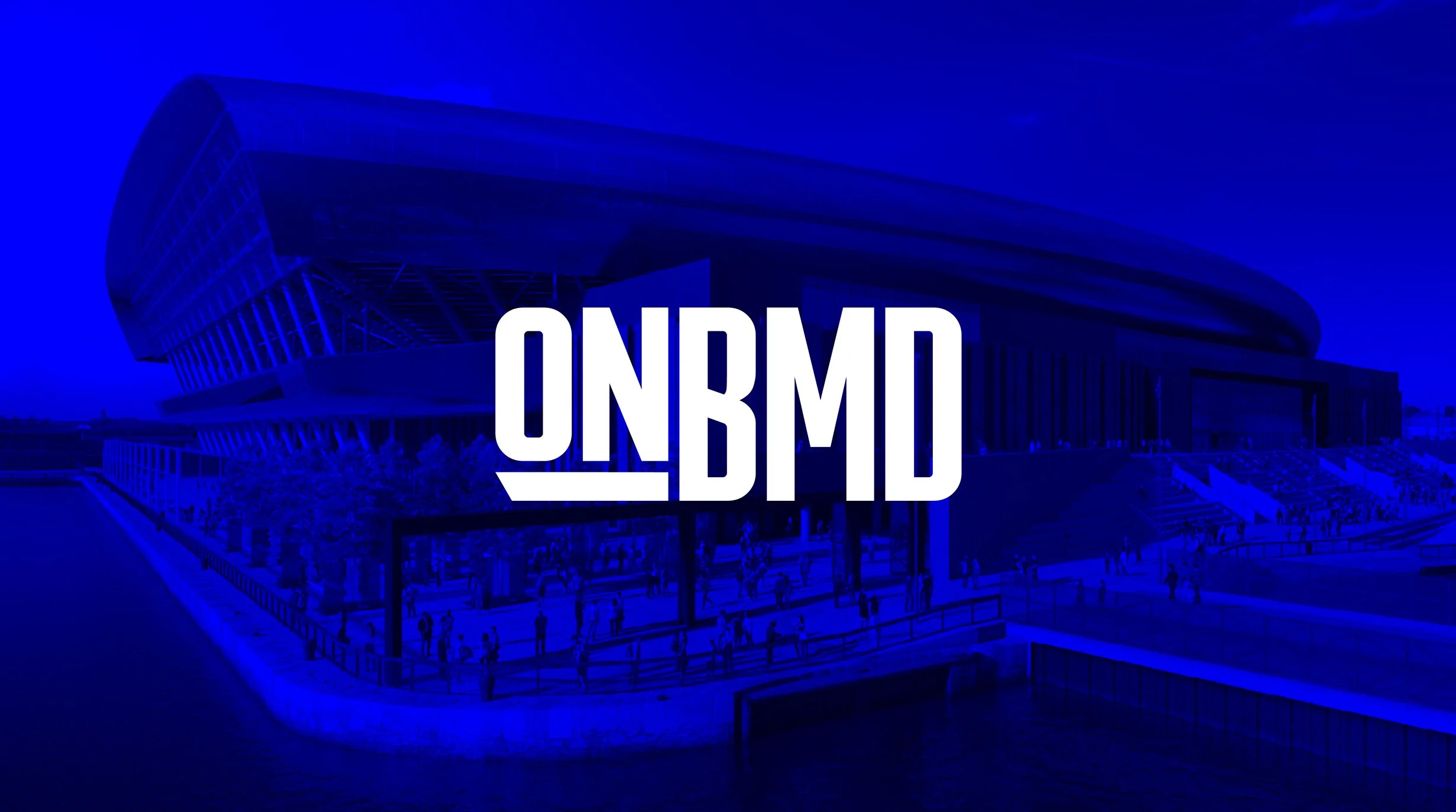 A modern stadium with a rounded roof and glass facade, with people walking outside and the letters ON3RMD in the center.