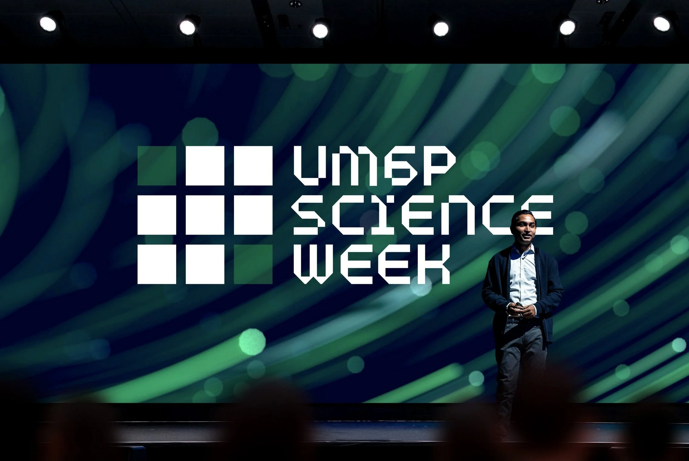 A man standing on a stage at Under Science Week event with a digital screen behind him displaying the event's logo with a grid of squares and the words 'Under Science Week'.