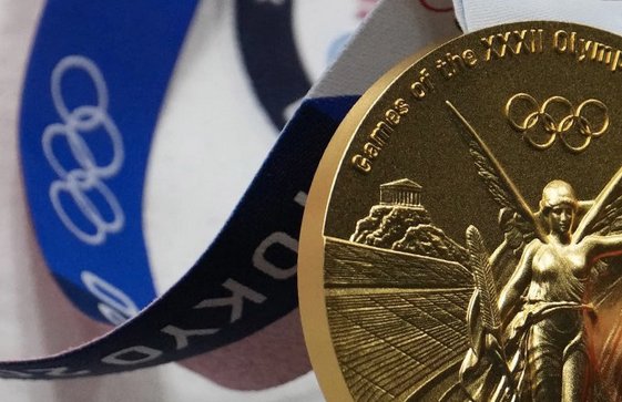 Close-up of a gold Olympic medal with a ribbon, depicting the goddess Nike holding a laurel wreath and swords, with part of the Olympic rings visible.
