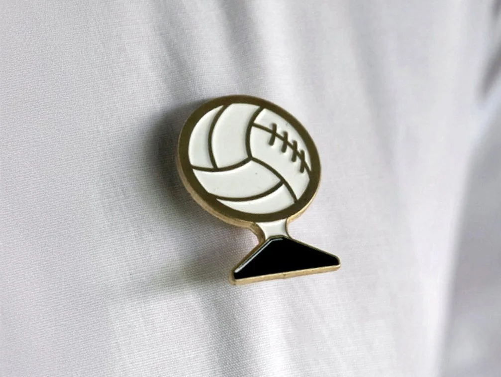 Pin featuring a volleyball on a stand, attached to a white fabric.