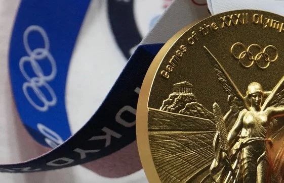 Close-up of a gold medal with the Olympic rings and a winged figure holding a torch, with a ribbon in the background.