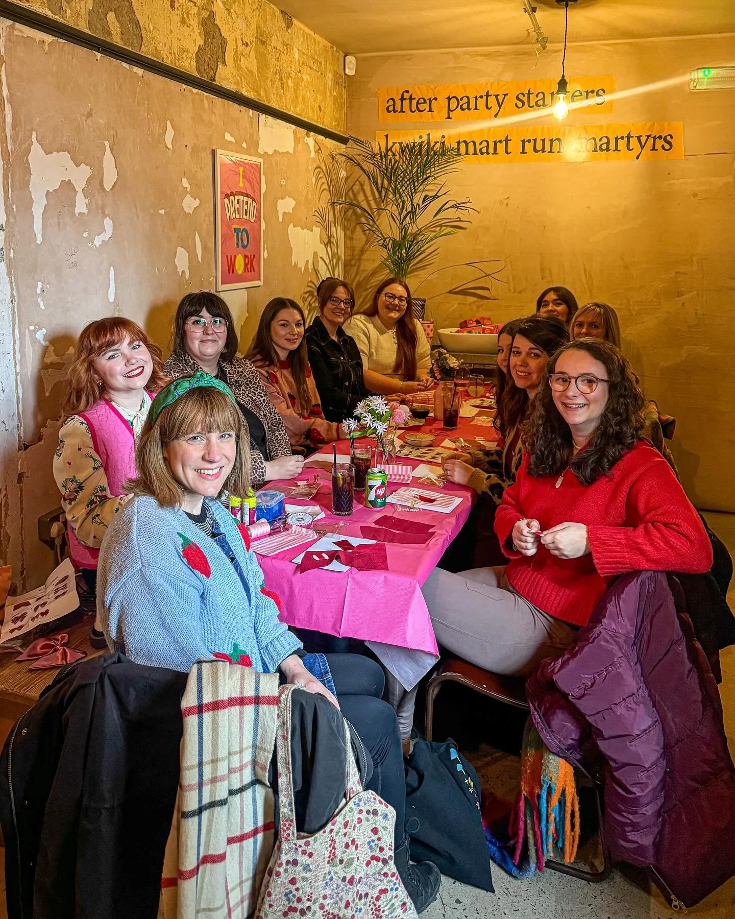 A group of women sitting around a pink tablecloth-covered table in a cozy, rustic restaurant, smiling at the camera. The background has a textured wall with a small poster that reads 'I Preaded to Work' and a sign that says 'after party starters knit 2,1 mart run martyrs.' There are drinks, flowers, and gift bags on the table, with a large potted plant in the corner.
