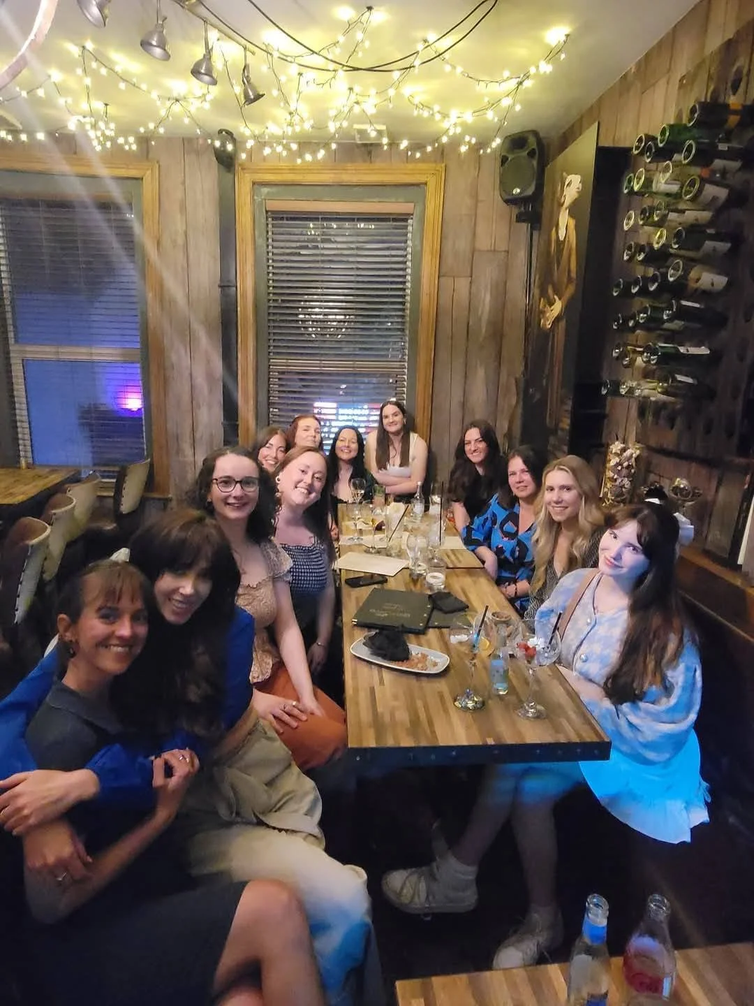 A group of women sitting around a wooden table in a cozy, warmly lit restaurant with string lights on the ceiling and wine bottles on the wall.