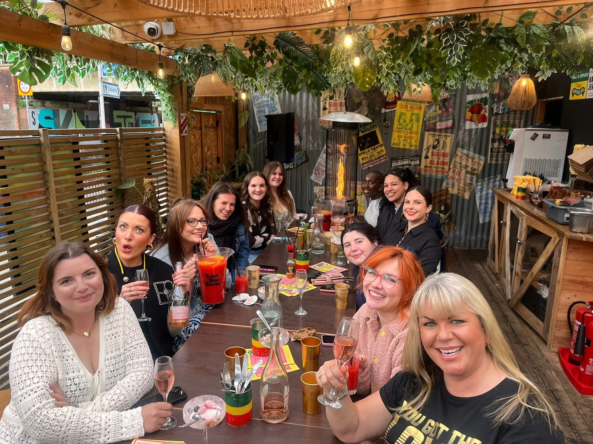 A group of women sitting around a long table at an outdoor or semi-outdoor venue, celebrating with drinks and smiling for the camera.