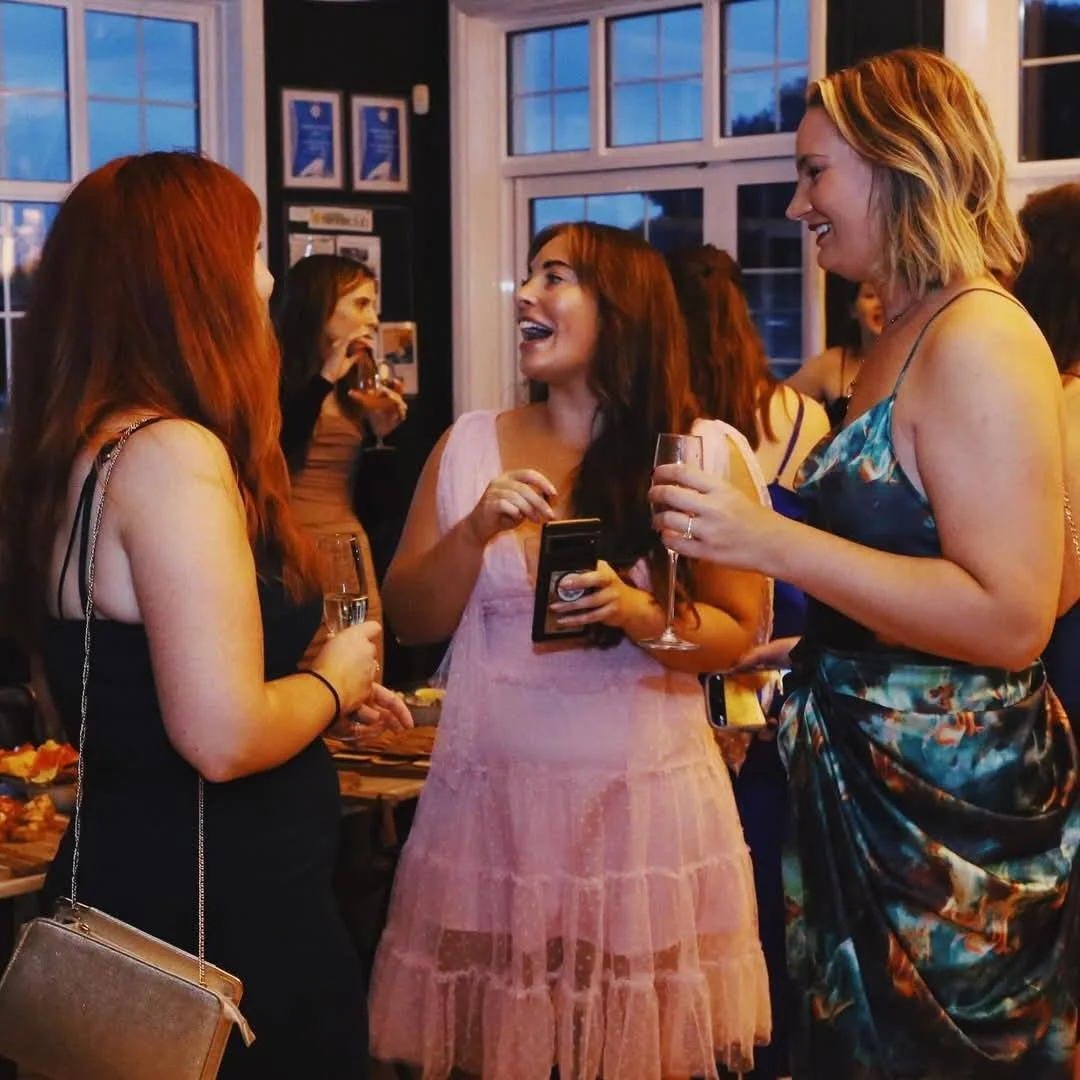 Three women sharing a laugh and holding glasses of wine at a social gathering in a warmly lit room with large windows.