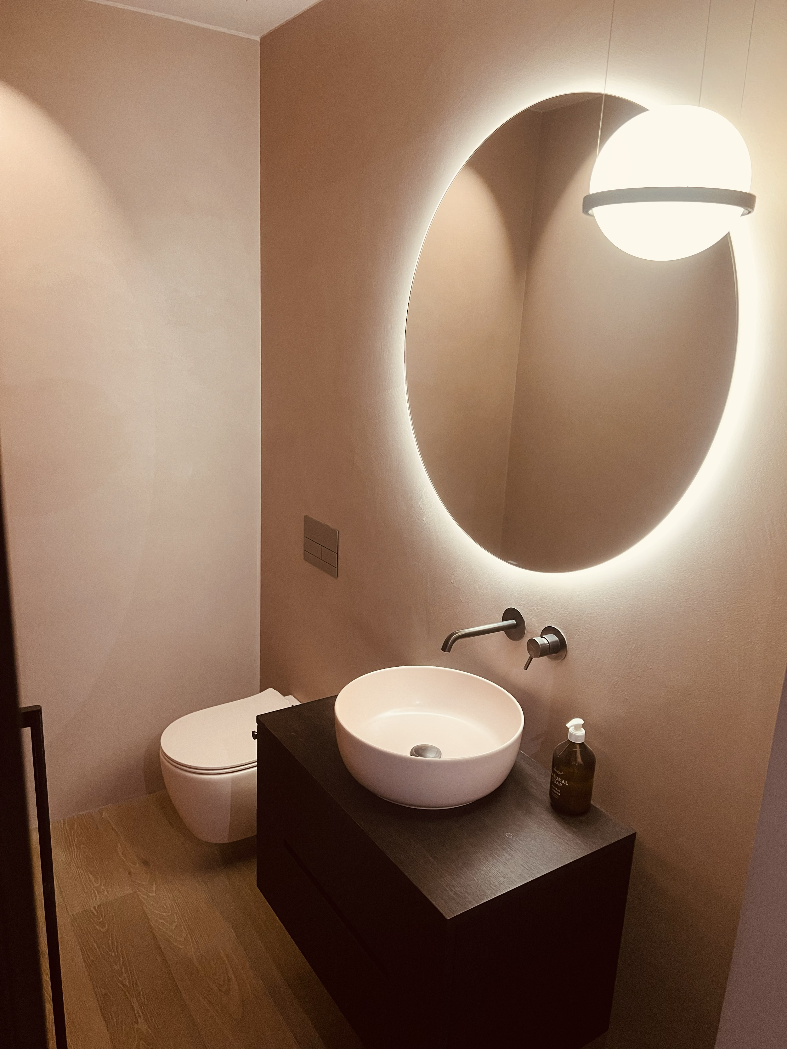 Moderner Badezimmerbereich mit wandhängender Toilette, schwarzem Waschtisch mit runder weißen Schüssel, großem ovalem Spiegel mit LED-Beleuchtung und angenehmer Beleuchtung durch Deckenlampe.