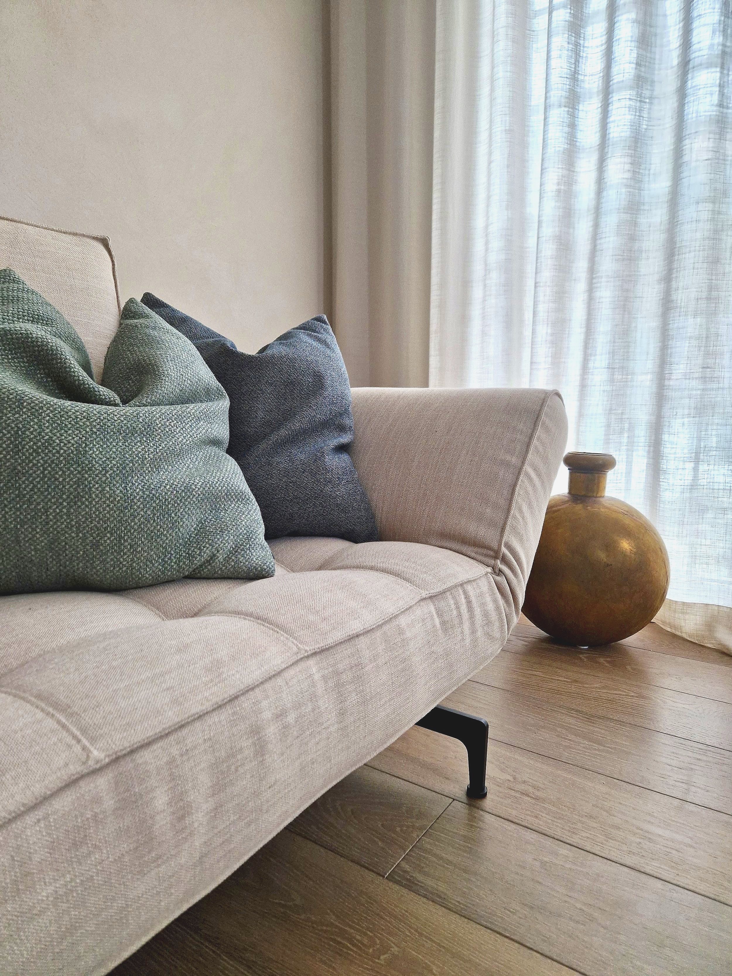 Wohnzimmer mit beige Couch, drei Kissen in Blau- und Grüntönen, goldene Vase auf Holzboden, vor weißen Vorhängen.