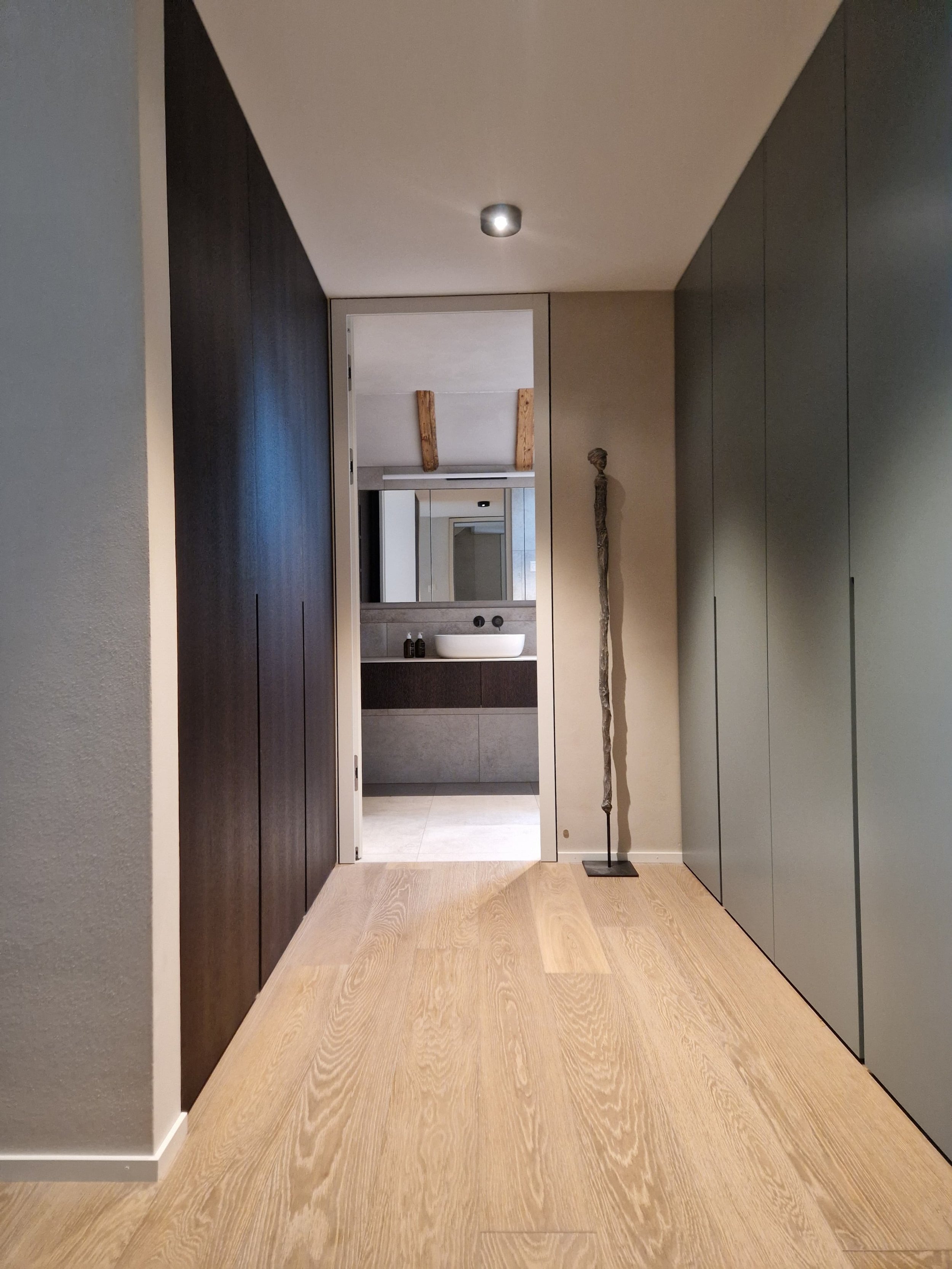Heller Flur mit Holzboden, dunklen und grauen Wänden, steinernem Skulptur und Blick ins Badezimmer mit Spiegel, Waschbecken und Holzregal