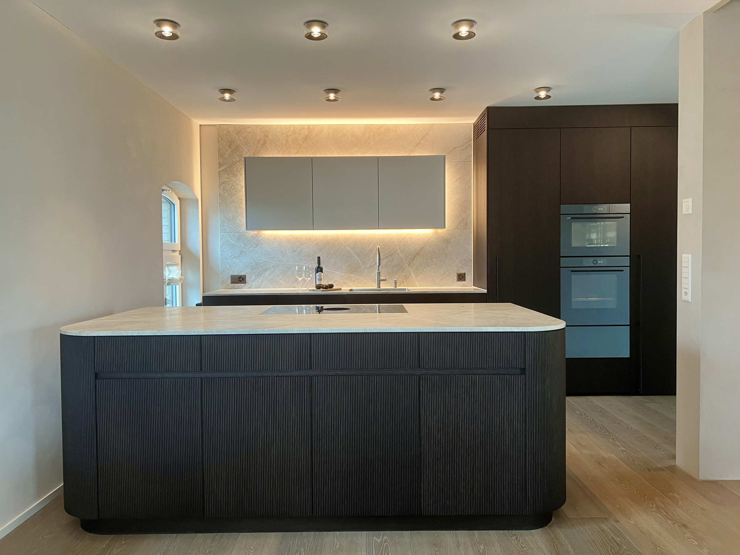 Moderne Küche mit dunklem Küchenblock, heller Arbeitsplatte, Backofen, Kochfeld, Wasserhahn, Weinflasche und zwei Gläser, beige Wand mit hintergrundbeleuchteten Schränken, kleine Fenster