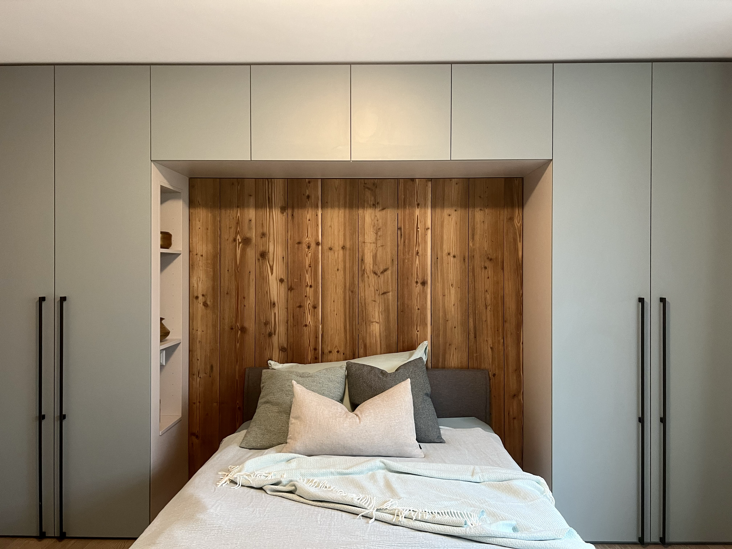 Bett mit grauen und weißen Kissen vor einer Holzvertäfelung, eingerahmt von weißen Schränken mit schwarzen Griffen