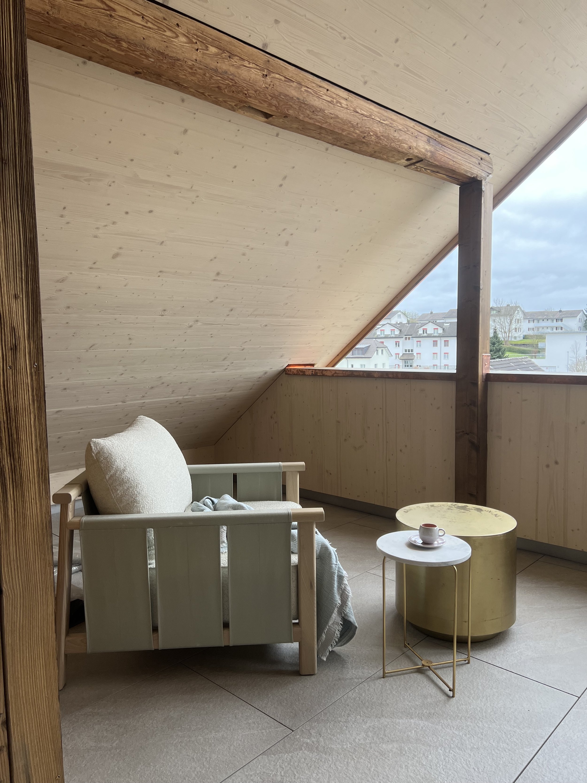 Ein gemütlicher Balkon mit einem kleinen weißen Sitzmöbel, zwei Beistelltischen aus Metall und Marmor, einer Tasse und einer Untertasse auf einem goldfarbenen Tisch. Der Balkon hat eine Dachschräge mit Holzvertäfelung und eine Aussicht auf eine Wohns